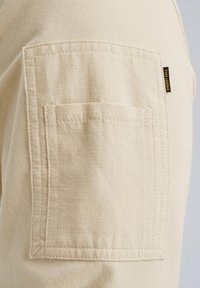 Beiger Baumwollstoff mit texturierter Oberfläche, ausgestattet mit einer quadratischen aufgenähten Tasche und einem gelben Etikett. Einfaches Design mit ordentlich genähten Kanten.