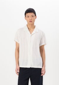 Camicia bianca a maniche corte con un leggero motivo a maglia testurizzato, dotata di colletto e chiusura a bottoni, abbinata a pantaloni scuri.