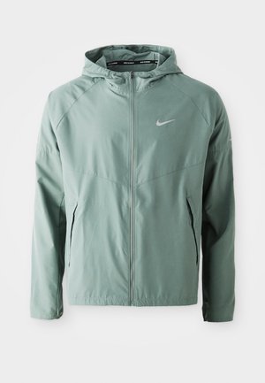 Jachetă Nike de culoare verde deschis, cu glugă, închidere cu fermoar, buzunare laterale și un logo reflectorizant. Fabricată dintr-un material neted, ușor.