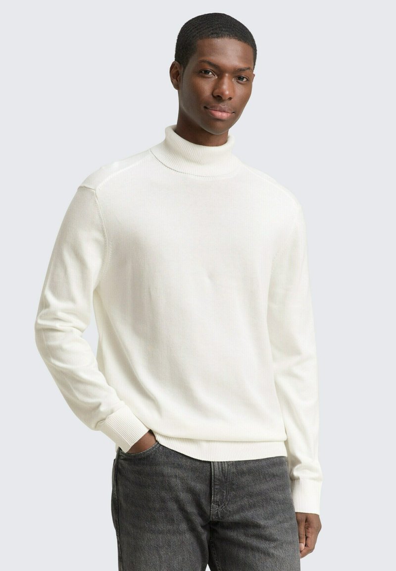 Weißer Rollkragenpullover mit geripptem Muster und langen Ärmeln, kombiniert mit dunkelgrauen Jeans. Das Model posiert mit einer Hand in der Tasche.