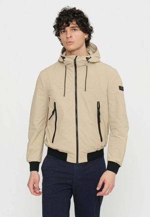 Giacca cappuccio beige chiaro con coulisse regolabile, zip nera e due tasche frontali; polsini ed orlo inferiore elasticizzati per una vestibilità aderente.