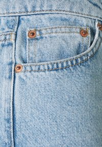 Vaqueros de mezclilla azul claro con herrajes de cobre, costuras visibles y un pequeño bolsillo trasero. La tela presenta un aspecto ligeramente desgastado y texturizado.