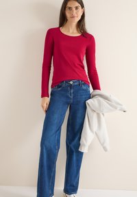 Maglietta rossa a maniche lunghe abbinata a jeans a gamba larga blu, mentre tiene una felpa grigia chiara. Tessuto morbido, vestibilità comoda e design semplice.