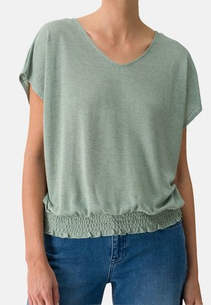 Haut vert clair à manches courtes avec un col en V et un ourlet froncé. Tissu doux et texturé avec une coupe décontractée, associé à un jean bleu.