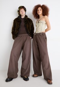 Braune weite Hosen mit elastischem Bund, kombiniert mit einem beigen trägerlosen Korsett und einer braunen Kunstfelljacke. Texturierte Schuhe sorgen für Details.