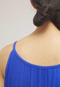 Top sans manches bleu avec un col froncé. Le tissu semble doux avec une texture subtile, révélant la peau dans le dos.