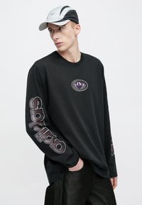 Juoda ilgomis rankovėmis marškinėliai su violetiniu logotipu ant krūtinės ir ryškiu, per rankoves einančiu „sp“ užrašu. Minkšta medžiaga ir laisvas siluetas.