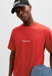 Punane puuvillane T-särk lühikeste varrukatega ja ümmarguse kaelusega, millel on rinnal valge tikandiga tekst "Originals Studio".