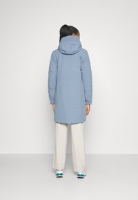 Parka impermeable de color azul claro con capucha, cuenta con una cintura ajustada, puños elásticos y un discreto detalle del logo en la parte posterior.