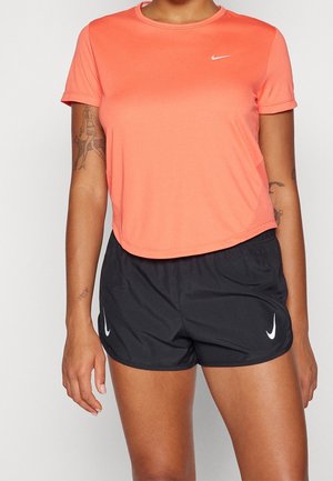 Personne portant un t-shirt Nike corail à manches courtes et un short de sport Nike noir, avec des tatouages visibles sur les deux bras.