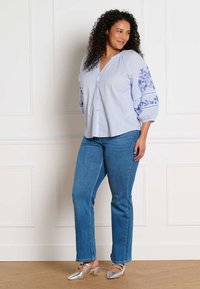 Femme aux cheveux bouclés, souriante, portant un chemisier bleu clair avec une broderie florale, un jean bleu et des chaussures argentées à petits talons.