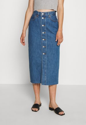 Kvinde iført en blå midi denim nederdel med knapper foran, frynset kant og sorte åbne slidesandaler mod en ensfarvet baggrund.