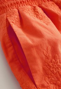 Oranger Stoff mit floraler Stickerei und gerafftem elastischem Bund, der die detaillierte Stichtextur und Falten zeigt.