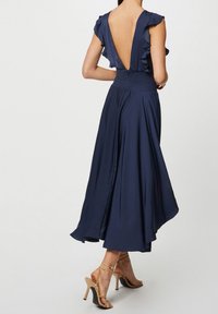 Kobieta w granatowej sukience midi z falbaniastymi rękawami i głębokim dekoltem na plecach, w połączeniu z beżowymi sandałami na wysokim obcasie.