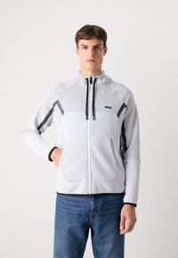 Veste zippée gris clair avec un haut texturé respirant, des rayures d'accent noires et un col haut. Comprend des poches avant et des poignets contrastants.