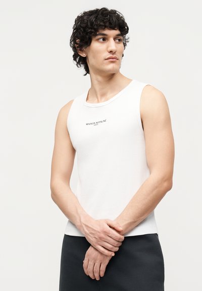 Νέος άνδρας με σγουρά σκουρόχρωμα μαλλιά που φοράει λευκό αμάνικο μπλουζάκι Maison Kitsuné, μαύρο παντελόνι, κοιτάει προς τα αριστερά με τα χέρια σταυρωμένα μπροστά.