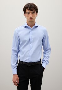 Joven vestido con una camisa de vestir rayada vertical en azul claro, metida en pantalones negros con un cinturón negro, de pie contra una pared lisa.