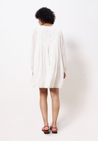 Weißes Leinenkleid mit langen Ärmeln, Raffung im Rücken und lockerer Passform, kombiniert mit roten Riemchensandalen.