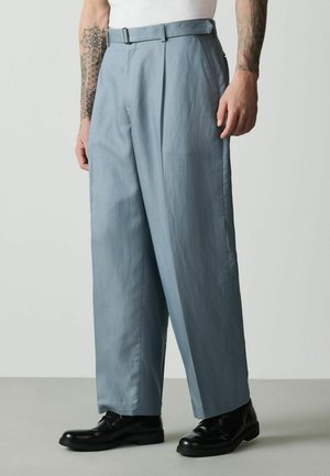 Mann trägt lockere hellblaue Hose, schwarze Anzugschuhe, weißes Hemd, mit Tätowierungen auf beiden Unterarmen vor einfarbigem Hintergrund.