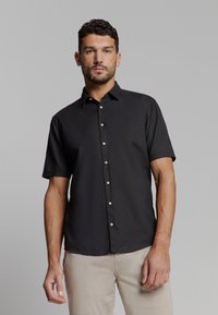 Zwart kortemmers overhemd met knoopsluiting en kraag, een voorpand met knopen en witte knopen. De stof lijkt textuur te hebben. Gemaakt met lichte broek.