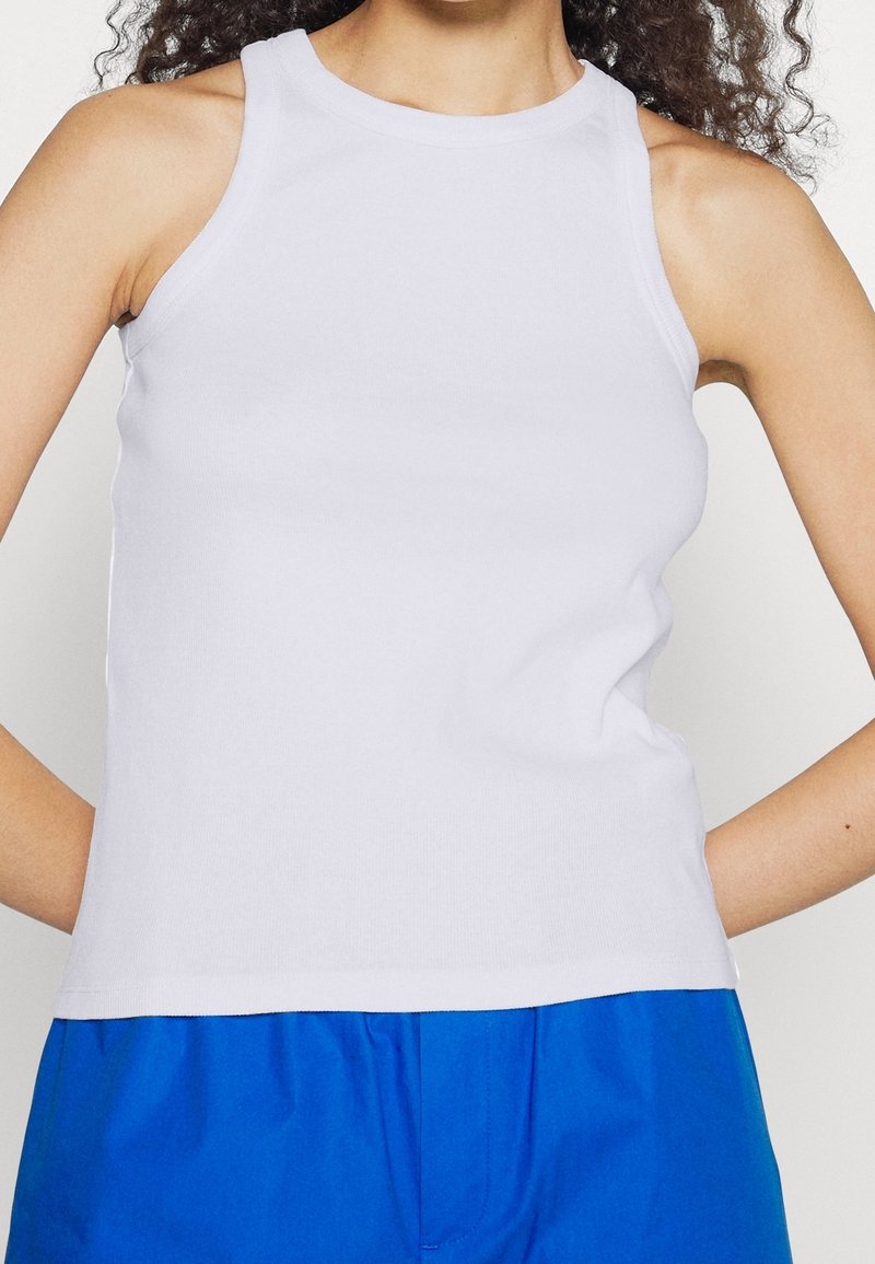 White sleeveless tank top with a high neckline, ribbed texture, and fitted design, paired with blue shorts in a casual style.