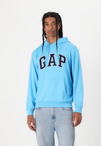 Light blue hoodie with a front pocket, featuring "GAP" in bold black letters. Made of soft fabric with a relaxed fit. Paired with light denim jeans.