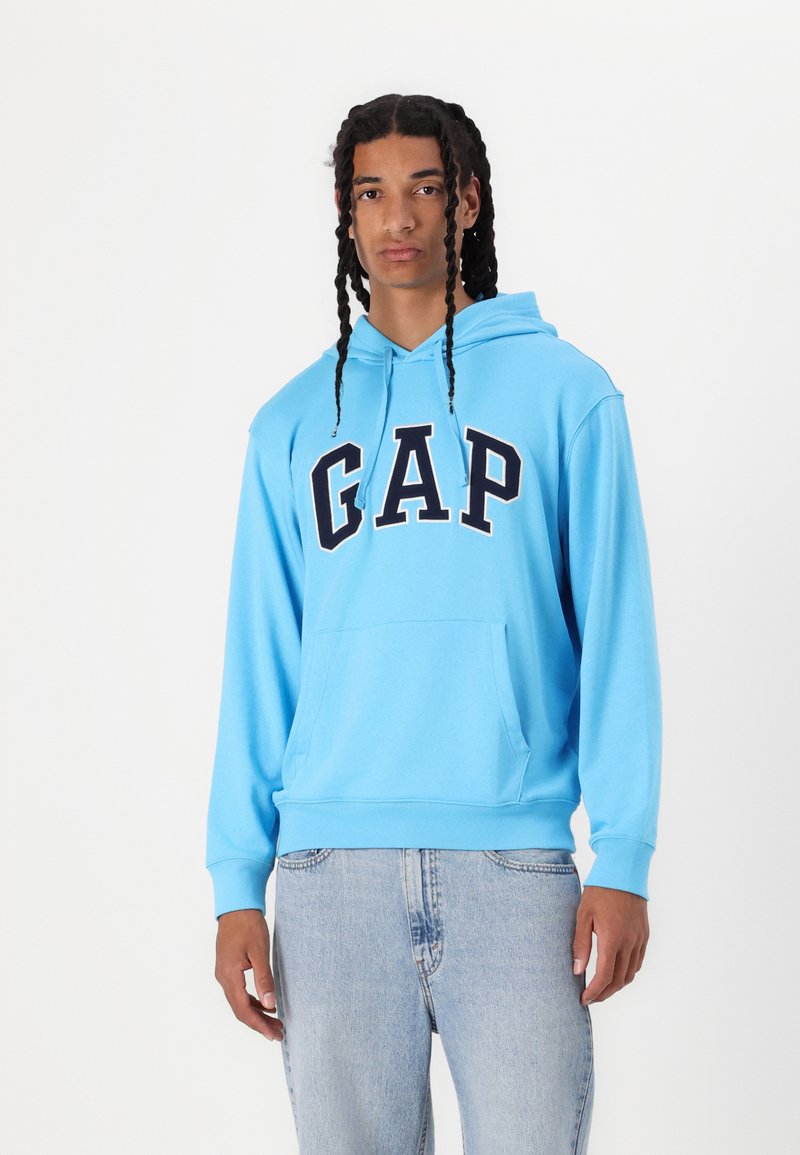 Light blue hoodie with a front pocket, featuring "GAP" in bold black letters. Made of soft fabric with a relaxed fit. Paired with light denim jeans.