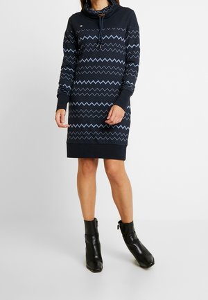 Femme portant une robe à manches longues bleu marine à motif zigzag, associée à des bottines noires, debout devant un fond blanc uni.
