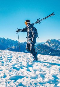 Skijder in donkere uitrusting draagt ski's over zijn schouder en houdt skistokken vast, staand op een besneeuwde berg met een heldere blauwe lucht en ruige toppen op de achtergrond.