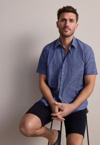 Lühikeste varrukatega, sinise lillemustriga särk kergefabrikus. Modell istub taburetil, seljas tumedad lühikesed püksid ja sandaalid.