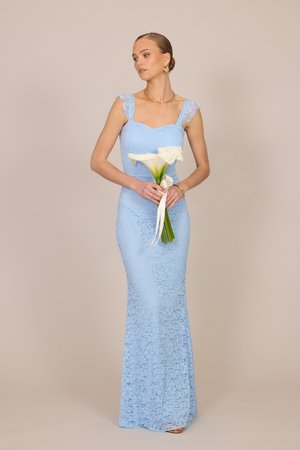 Femeie într-o rochie albastru deschis din dantelă fără mâneci, ținând un buchet de cale albe cu un fundal bej neutru.
