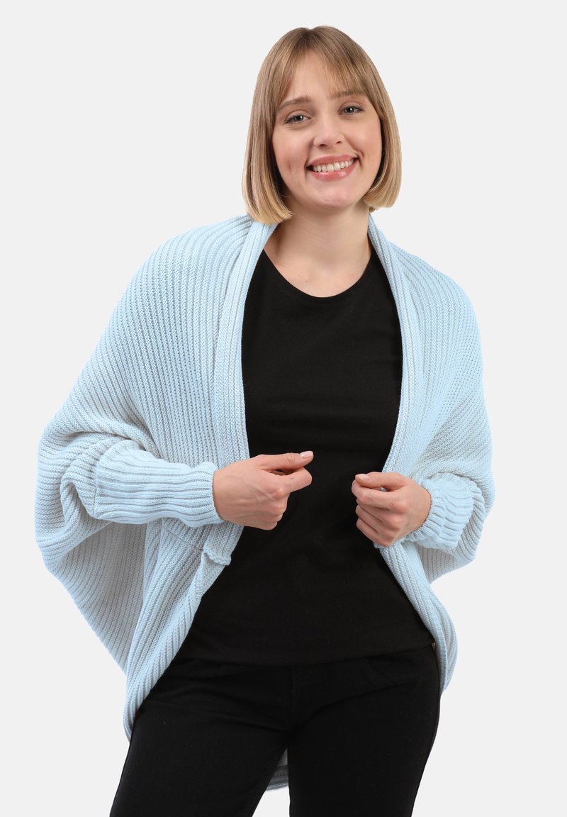 Cardigan a maglia azzurro chiaro con texture a coste, apertura frontale e maniche a campana. Indossato sopra una maglietta nera, evidenziando un look stratificato casual.