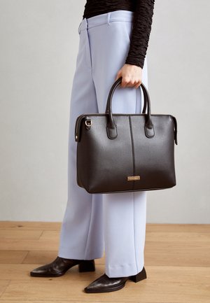 Person i lyseblå bukser og sort top, der holder en tekstureret sort lædertaske med dobbelt håndtag mod en ensfarvet baggrund.