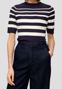 Kvinna som bär en marinblå och vit horisontellt randig kortärmad topp instoppad i mörka högmidjade jeans med ena handen i fickan.