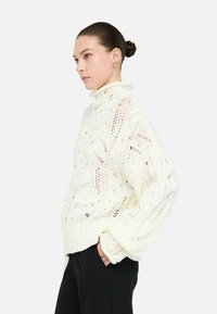 Donna con capelli raccolti a chignon che indossa un maglione bianco lavorato a maglia con decorazioni di perline e pantaloni neri, in piedi con le mani in tasca, rivolta a sinistra.