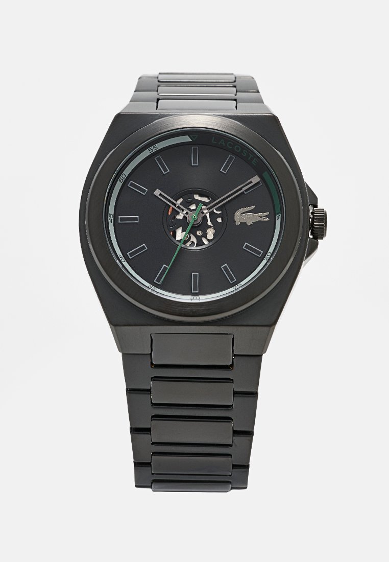 Reloj de pulsera de acero inoxidable negro con esfera redonda, engranajes internos visibles, marcadores de hora plateados, segundero verde y logo del cocodrilo de Lacoste.