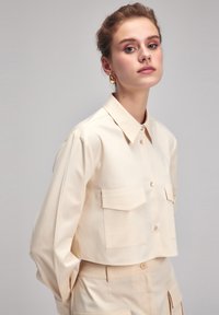 Korte crèmekleurige shirt van gladde stof met knoopsluiting aan de voorkant, twee borstzakken en een klassieke kraag. Heeft een verlaagde schouderlijn.