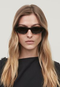 Occhiali da sole neri con un design rettangolare elegante e lenti scure. Montatura minimalista con texture liscia, indossati con una maglietta scura.