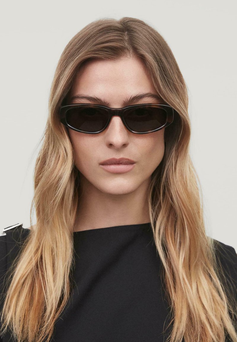 Occhiali da sole neri con un design rettangolare elegante e lenti scure. Montatura minimalista con texture liscia, indossati con una maglietta scura.