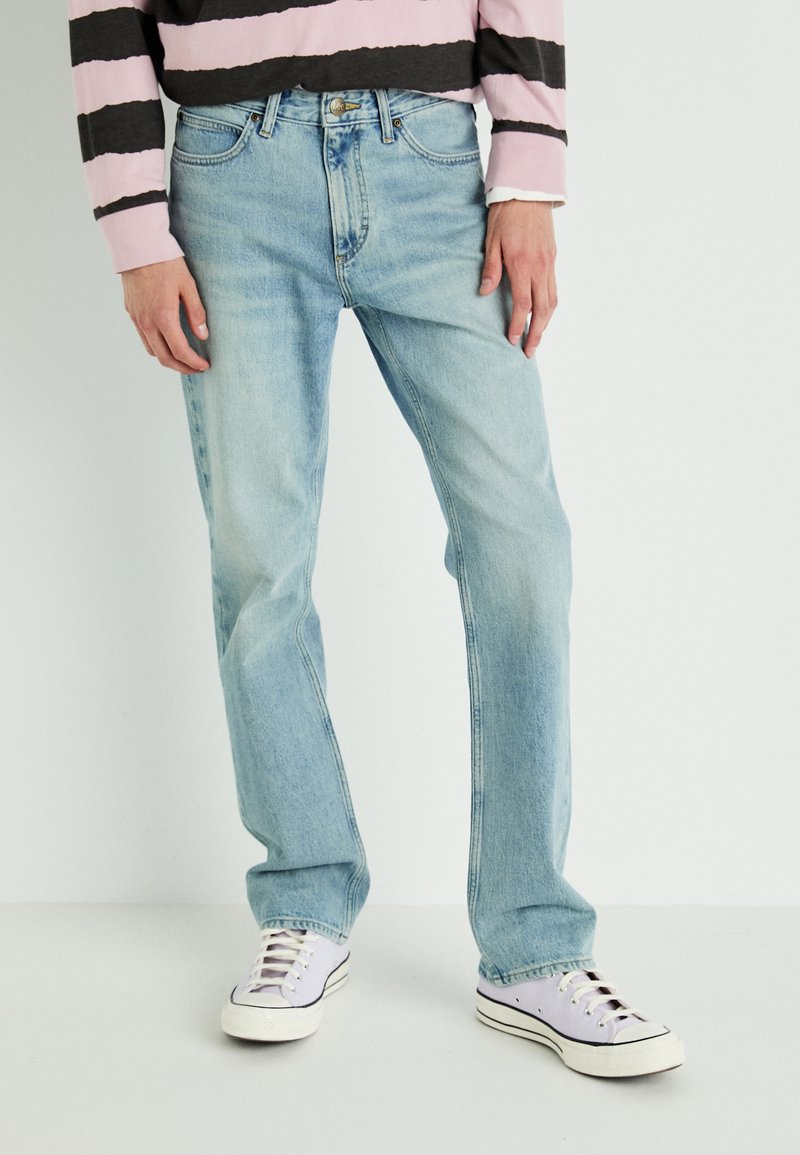 Jeans de denim azul claro con un corte relajado, que presentan un diseño tradicional de cinco bolsillos y un sutil desvanecimiento, combinados con zapatillas blancas.