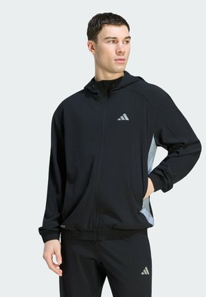 Homme portant une veste à capuche noire zippée et un pantalon assorti avec des panneaux gris clair et des logos, debout, les mains dans les poches, regardant vers la droite.