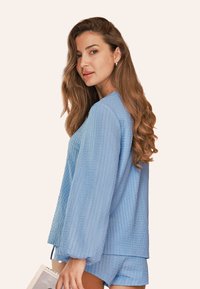 Blouse bleu clair texturé avec des manches longues et bouffantes et une coupe décontractée, associée à un short coordonné, portée par un mannequin sur un fond clair.