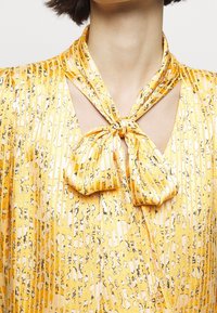 Top jaune enroulé avec un ruban noué au niveau du décolleté, des rayures verticales et un tissu texturé avec des motifs abstraits noirs.