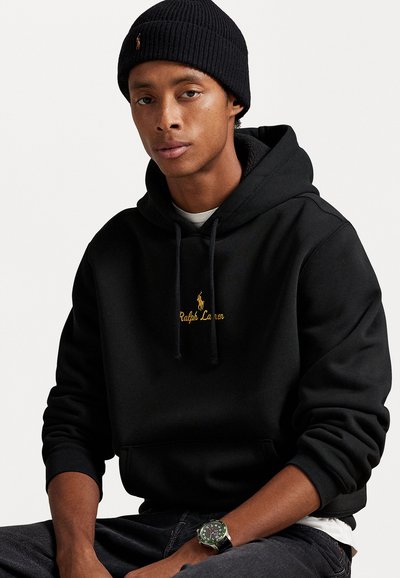 Vīrietis ar melnu adītu cepuri, melnu Ralph Lauren kapuci, tumšām džinsām un rokas pulksteni, sēž pret vienkāršu baltu fona.