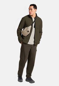 Olivgrüne Fleecejacke mit Reißverschluss vorne, kombiniert mit beigen Cargo-Hosen und einer beigen Umhängetasche aus strukturiertem Stoff.