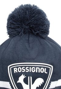 Námořnická pletená čepice s texturovaným povrchem, vybavená velkým huňatým bambulím nahoře a bílým logem kohouta v erbu.