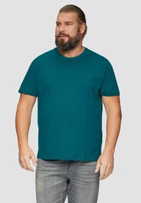 Turkusowa koszulka z krótkim rękawem wykonana z bawełny, z okrągłym dekoltem i małym logo na klatce piersiowej. Noszona z szarymi dżinsami.