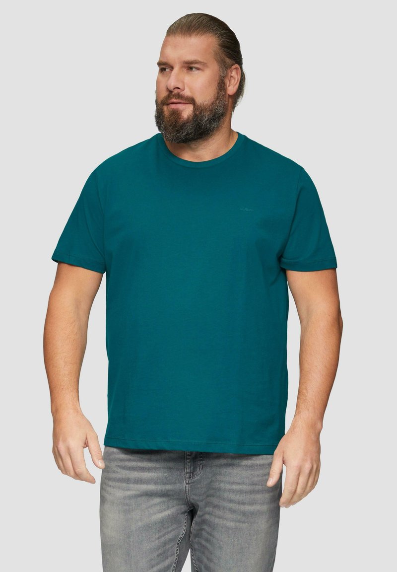 Turkusowa koszulka z krótkim rękawem wykonana z bawełny, z okrągłym dekoltem i małym logo na klatce piersiowej. Noszona z szarymi dżinsami.