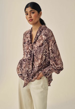 Bruine en beige blouse met slangprint, voorzien van een gedrapeerde kraag en losse mouwen, gecombineerd met lichtgekleurde broek, waarbij de gladde stoftextuur zichtbaar is.