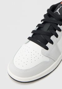 Λευκό δερμάτινο αθλητικό παπούτσι με γκρι λεπτομέρειες, διάτρητη μύτη, μαύρα κορδόνια και μαύρο-κόκκινο λογότυπο στη γλώσσα. Λουστραρισμένη ελαστική σόλα.